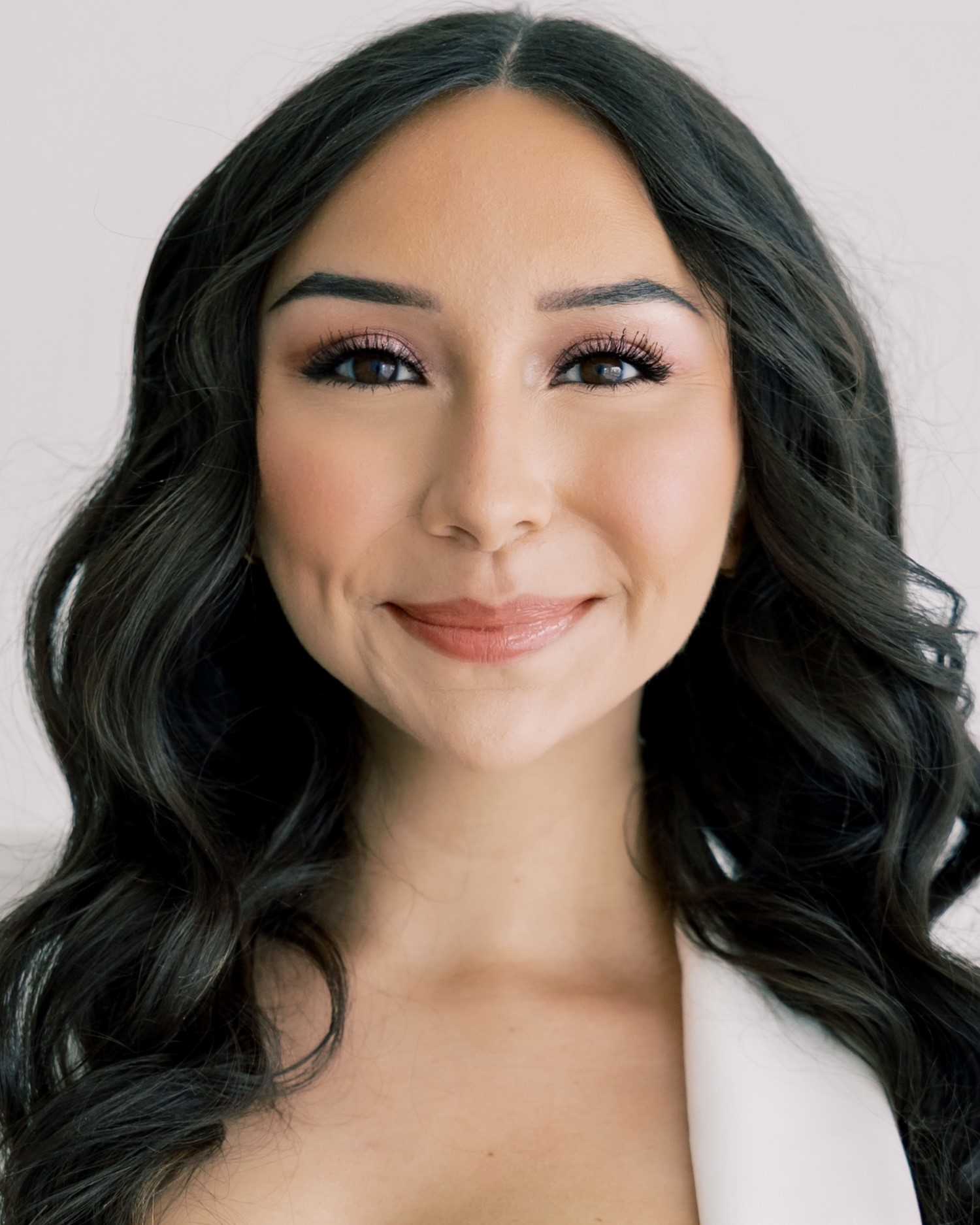 A young woman with long black hair smiles as she poses for a professional portrait. She is wearing a white jacket and light-red lipstick.