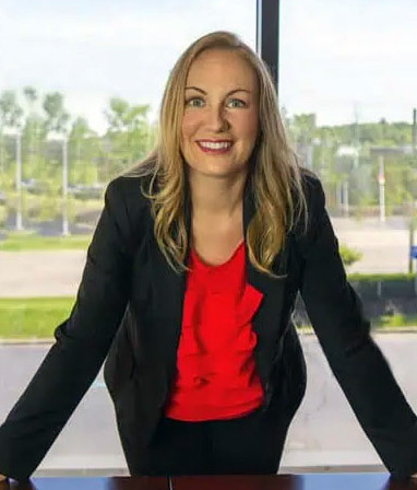 A person in a black blazer and red top stands with their hands on a table, looking out a large window with greenery and a parking lot visible outside.