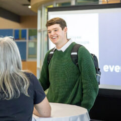 A person wearing a green sweater and carrying a backpack stands at a table, engaging in conversation with another individual. In the background, a screen displays the General Motors logo and text related to employee groups.