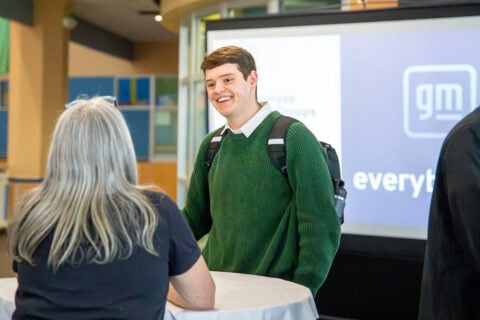 A person wearing a green sweater and carrying a backpack stands at a table, engaging in conversation with another individual. In the background, a screen displays the General Motors logo and text related to employee groups.