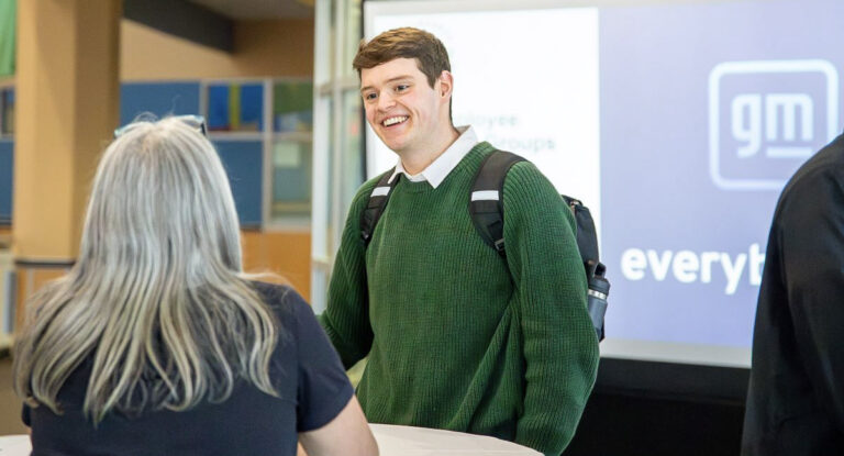 A person wearing a green sweater and carrying a backpack stands at a table, engaging in conversation with another individual. In the background, a screen displays the General Motors logo and text related to employee groups.
