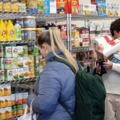 A person is selecting items from a shelf stocked with various canned goods, condiments, and packaged foods in a store. Another person is holding several items, including paper towels and boxes, while looking at the shelves.