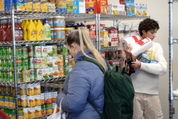 A person is selecting items from a shelf stocked with various canned goods, condiments, and packaged foods in a store. Another person is holding several items, including paper towels and boxes, while looking at the shelves.