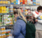 A person is selecting items from a shelf stocked with various canned goods, condiments, and packaged foods in a store. Another person is holding several items, including paper towels and boxes, while looking at the shelves.