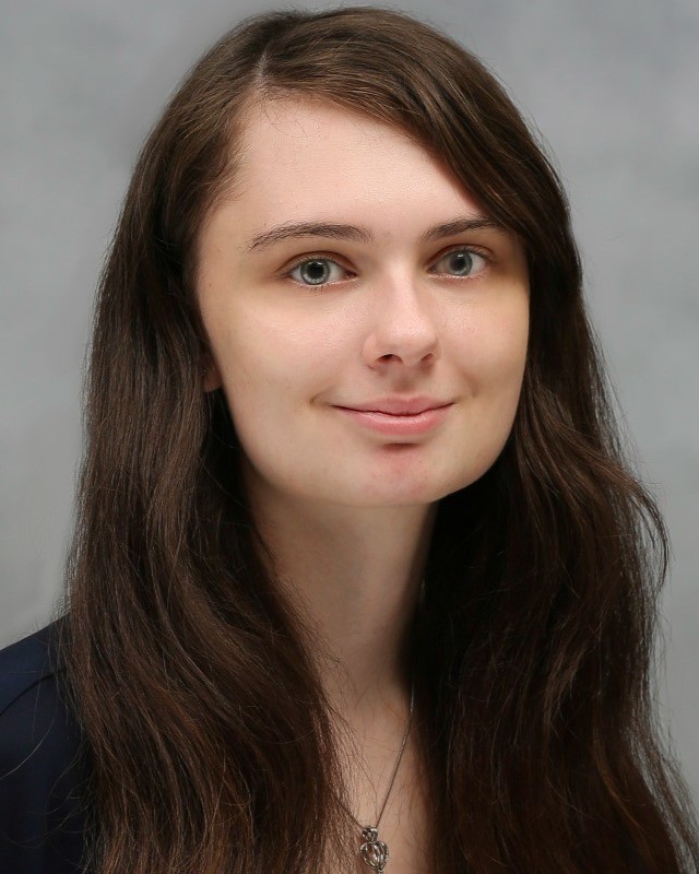 A young woman with long, wavy brown hair parted to the side looks directly at the camera with a gentle smile. She has light skin and blue eyes, and she is wearing a dark-blue top and a silver necklace with a small pendant.