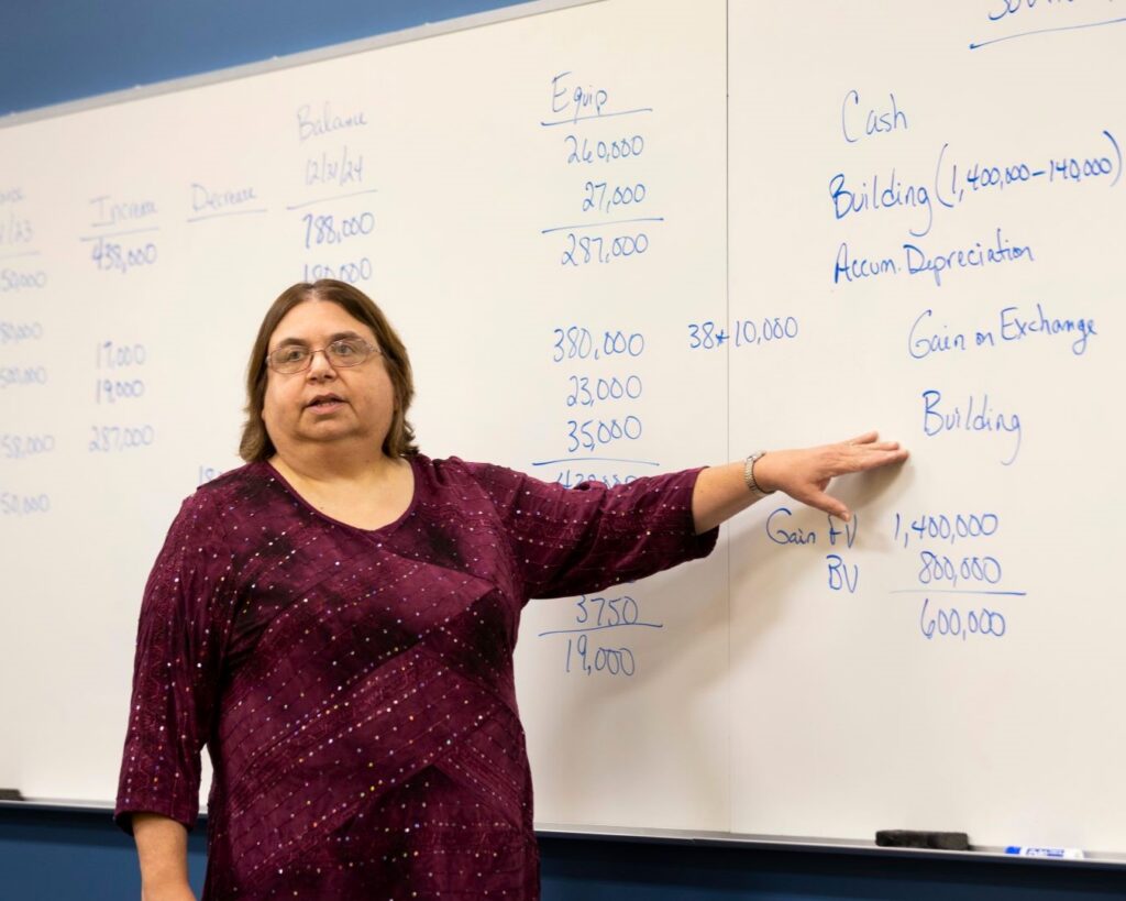 A female instructor stands in front of a large whiteboard filled with numbers and accounting terms, pointing toward the notes as she speaks.