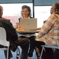 A woman smiles with her laptop in front of her. She is sitting across a table from a couple inside a meeting room with large windows at her back.