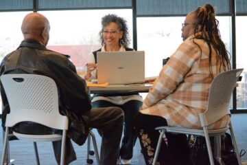 A woman smiles with her laptop in front of her. She is sitting across a table from a couple inside a meeting room with large windows at her back.