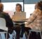 A woman smiles with her laptop in front of her. She is sitting across a table from a couple inside a meeting room with large windows at her back.