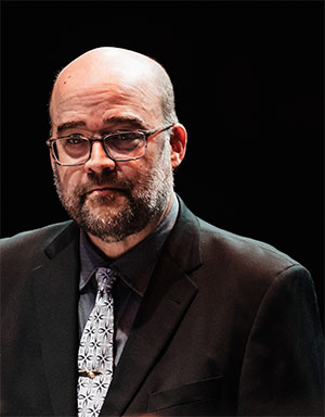 A man in a black suit and patterned tie stands against a dark background.