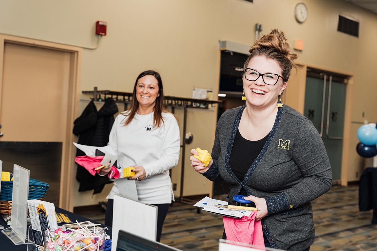 Two women at an indoor event table holding promotional items and papers, both wearing University of Michigan apparel.