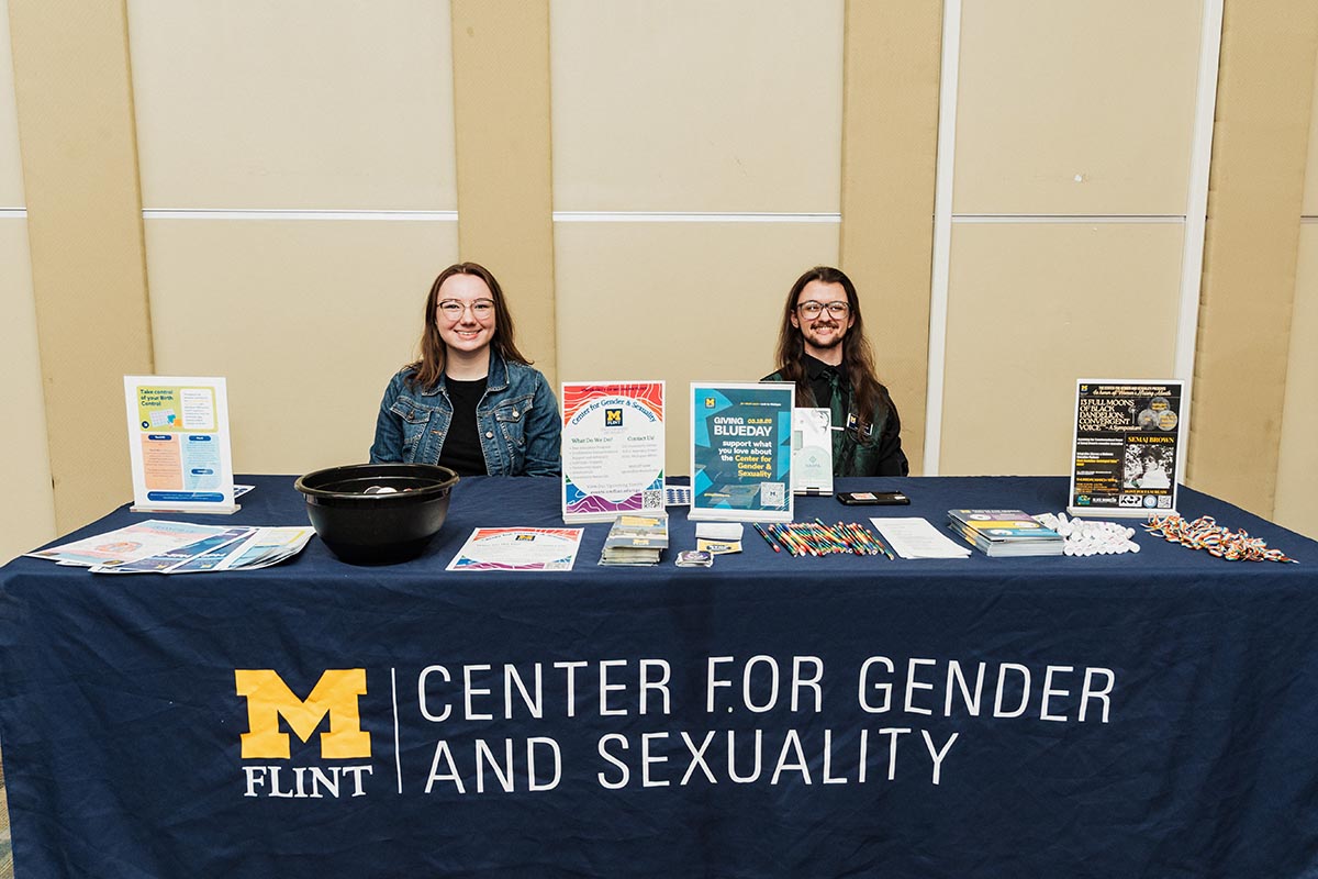 Two people sitting behind a table with a blue tablecloth that reads "M Flint Center for Gender and Sexuality," with informational materials and promotional items displayed on the table.