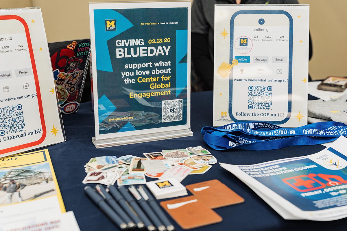 Table display with University of Michigan Flint Center for Global Engagement promotional materials including signs for Giving Blue Day on 03.18.26, QR codes for Instagram, pens, stickers, lanyards, and informational brochures.