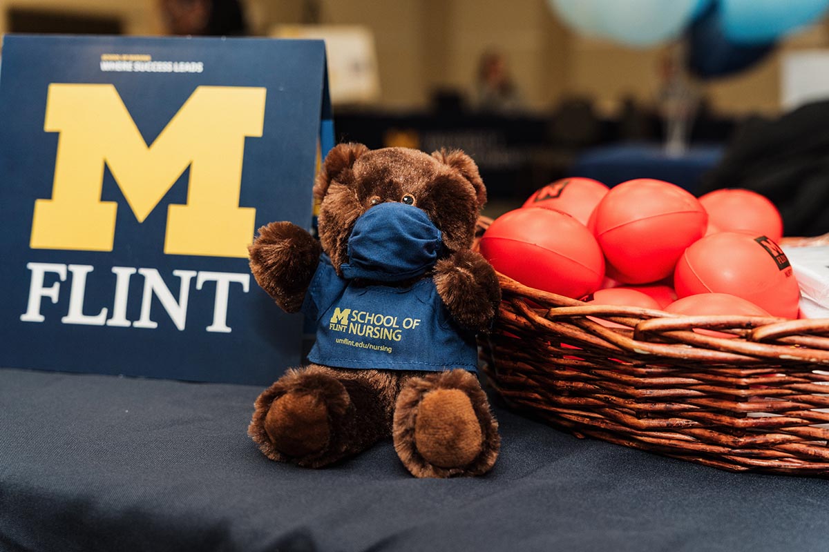 A brown teddy bear wearing a blue mask and a blue shirt with "M Flint School of Nursing" printed on it, sitting next to a basket of red stress balls and a sign with a large yellow "M" and the word "Flint."