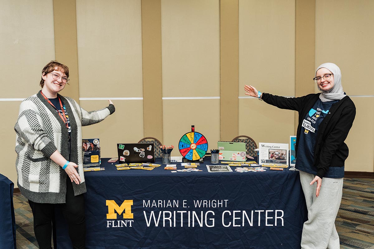 Two people stand on either side of a table with a blue tablecloth that reads "Marian E. Wright Writing Center" with the University of Michigan-Flint logo, presenting the table with various promotional materials and a colorful prize wheel.
