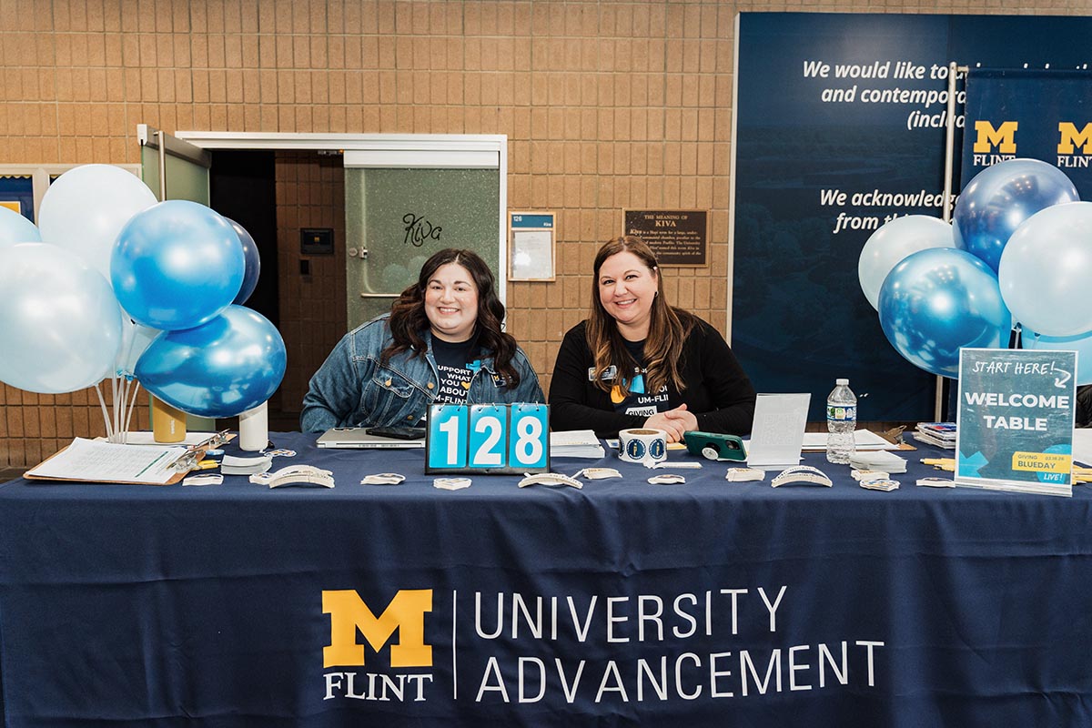 Two people sitting behind a University of Michigan Flint Advancement welcome table decorated with blue and white balloons and a sign that says "Start Here! Welcome Table."