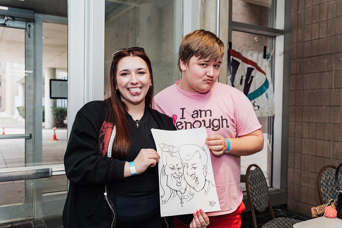 Two people holding a caricature drawing of themselves indoors near a glass door.