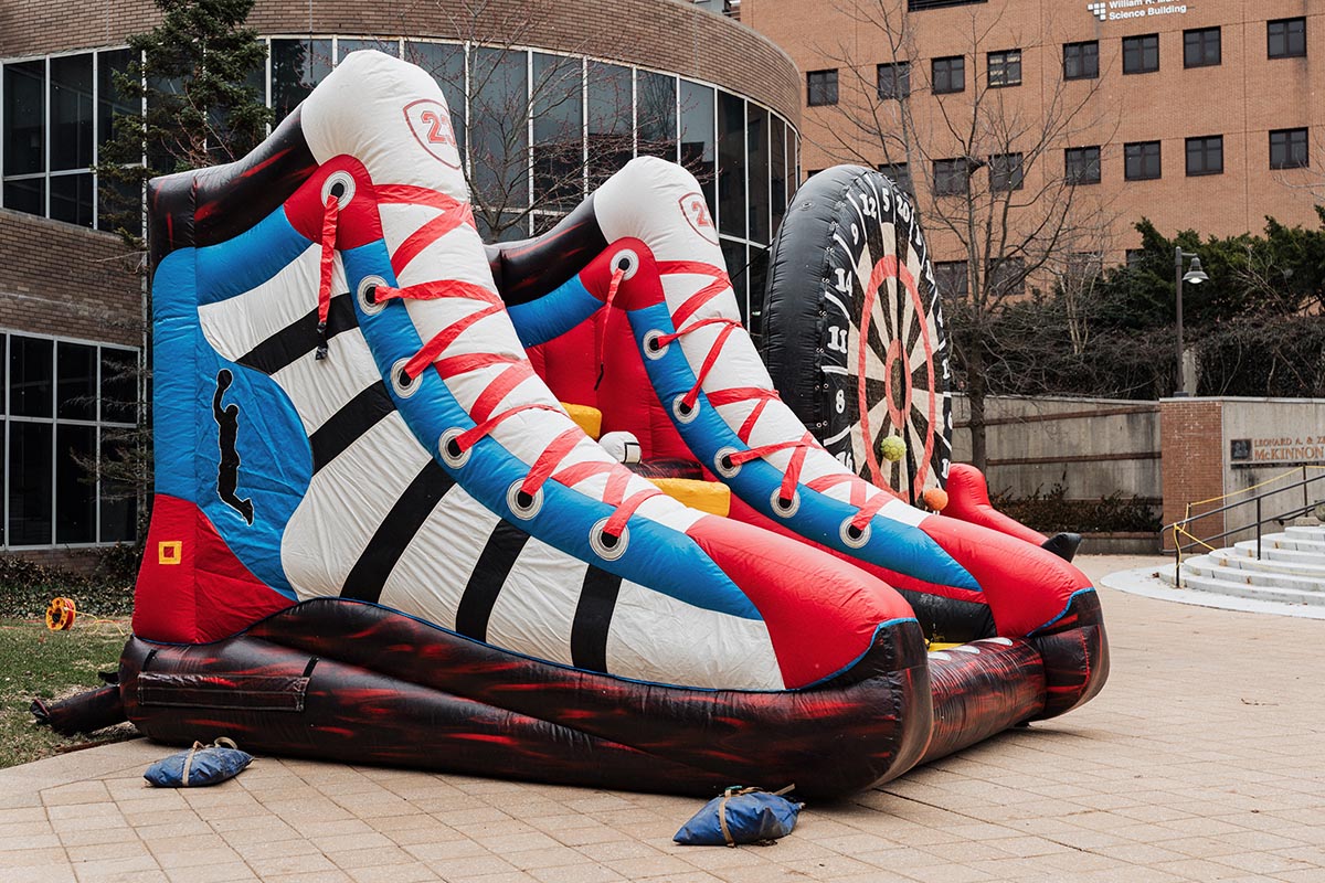 Inflatable game shaped like two large sneakers with a dartboard at the back, set up outdoors near a building.