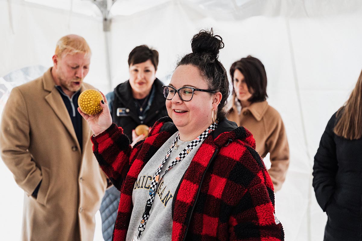 A person wearing a red and black checkered jacket and a gray Michigan sweatshirt is holding a yellow textured ball, with three other people standing in the background.