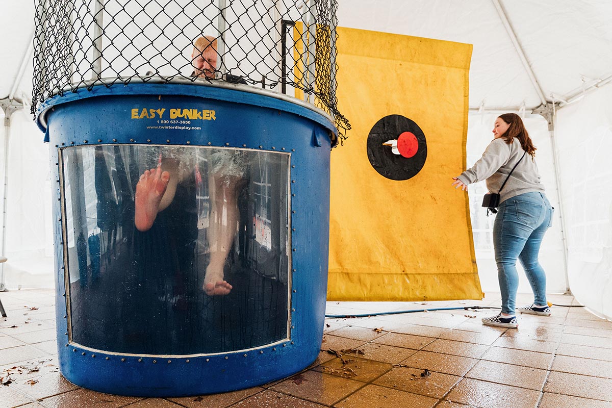 Person sitting inside a blue dunk tank with feet visible through a clear panel, while another person hits the target