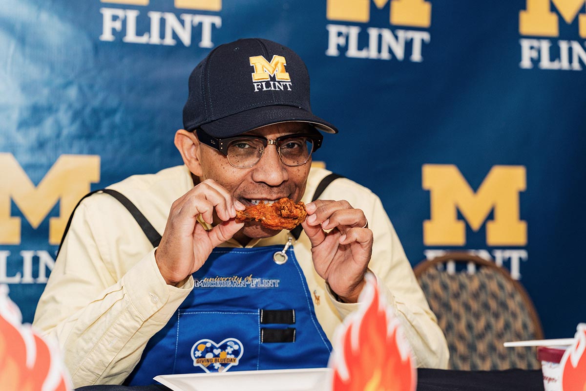 Chancellor Alexander wearing a University of Michigan-Flint cap and apron, eating a hot wing, with a blue University of Michigan-Flint backdrop behind them.