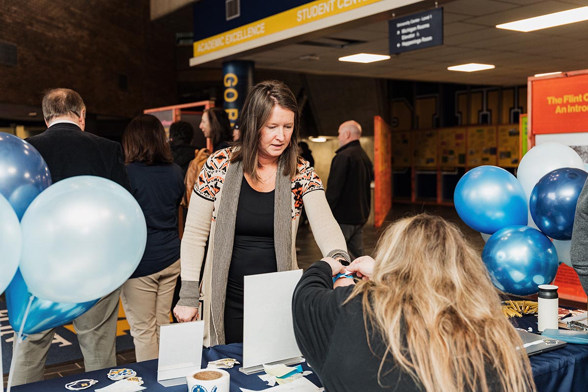 A woman is receiving a wristband from another woman at a table decorated with blue and white balloons in a student center.