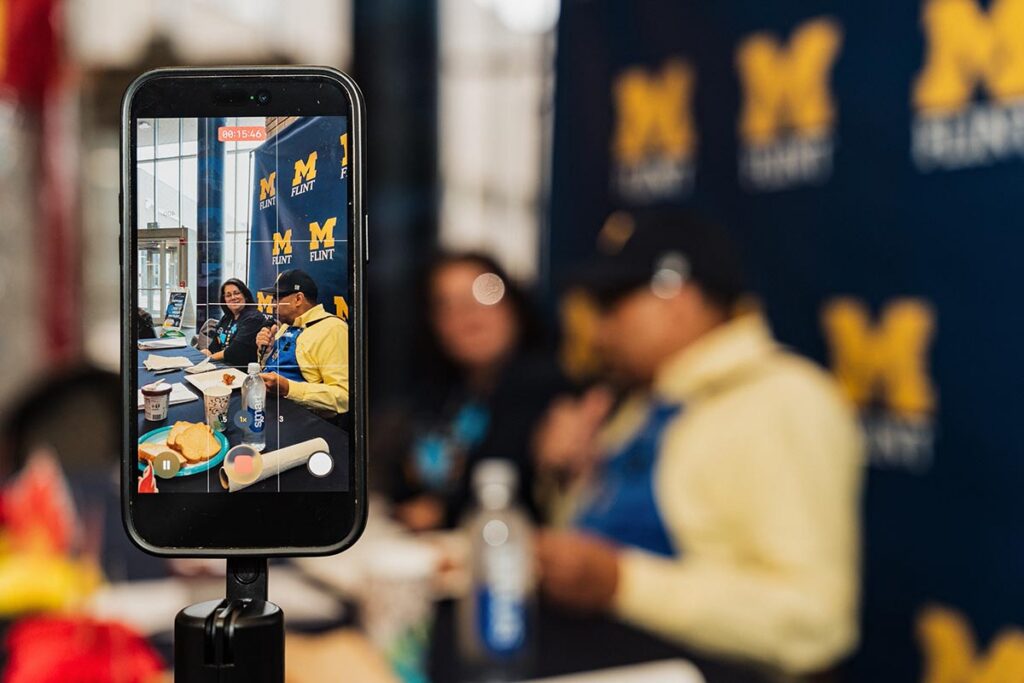Smartphone on a tripod recording two people sitting at a table with Michigan Flint banners in the background.