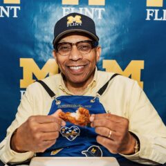 Chancellor Alexander wearing a Michigan Flint cap and a blue apron holding a hot wing in front of a Michigan Flint backdrop.