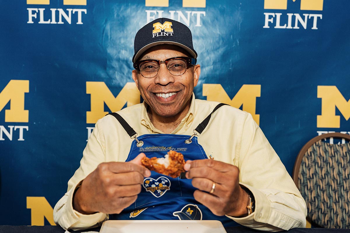 Chancellor Alexander wearing a Michigan Flint cap and a blue apron holding a hot wing in front of a Michigan Flint backdrop.