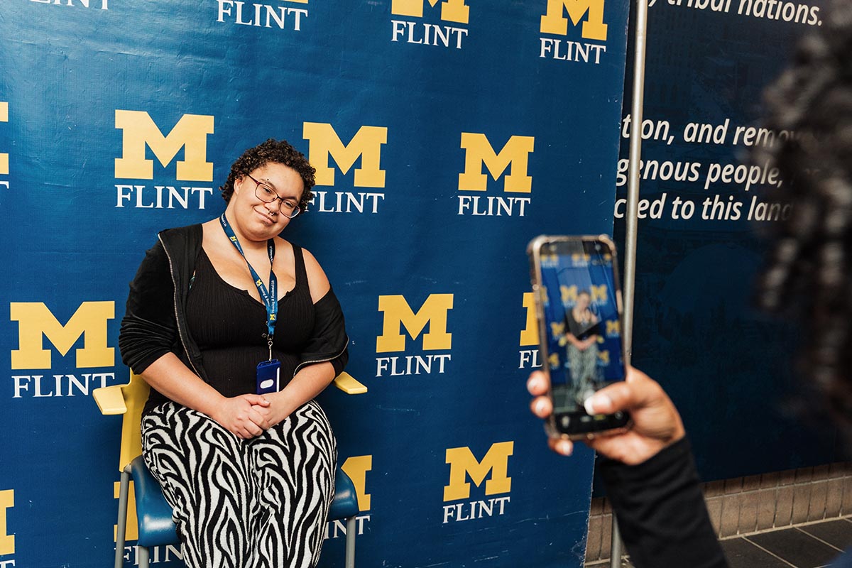 Person sitting in front of a blue backdrop with yellow "M Flint" logos while another person takes their photo with a smartphone.