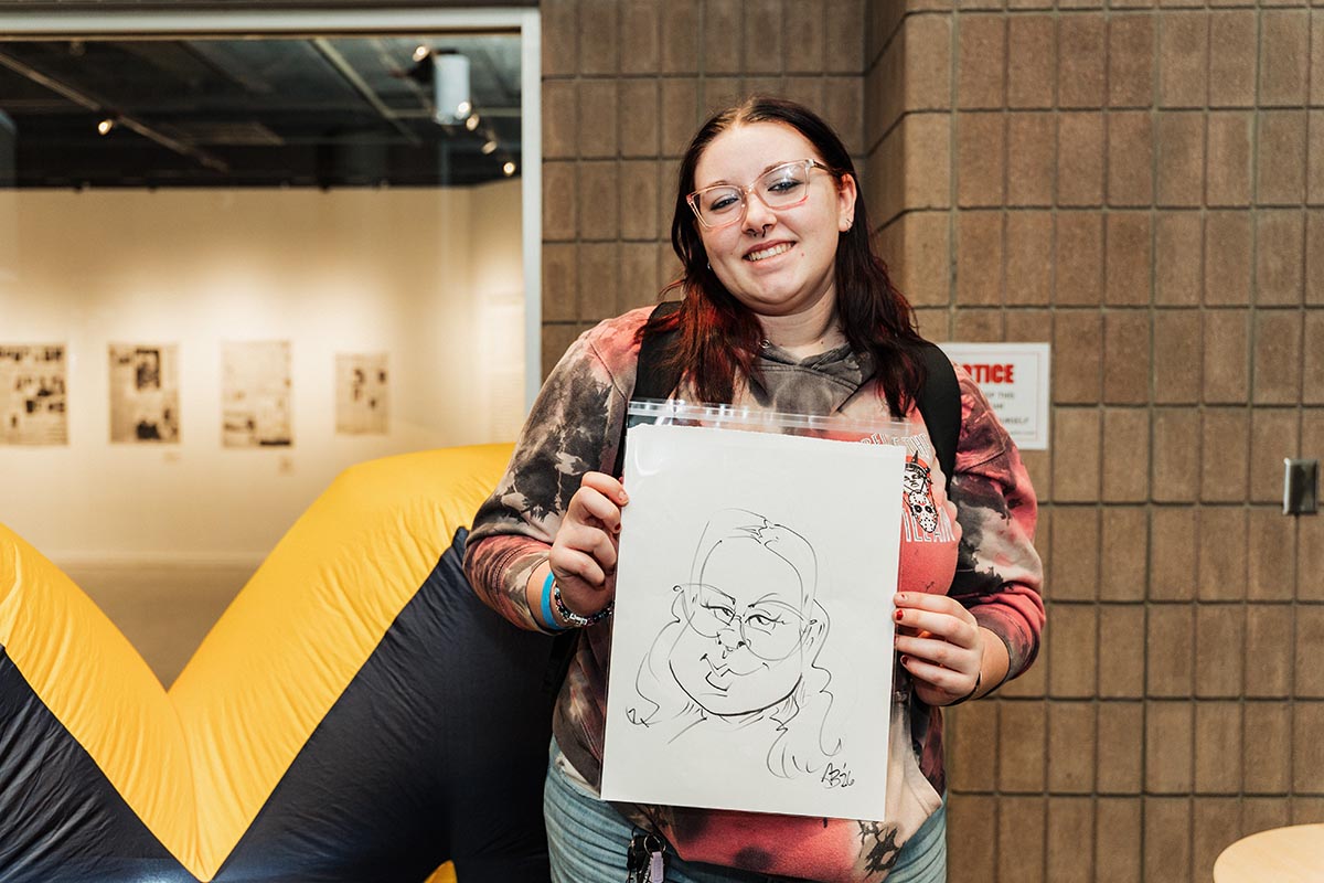 Person wearing a tie-dye hoodie and jeans holding a caricature drawing in an indoor setting with a yellow and black inflatable structure behind them.