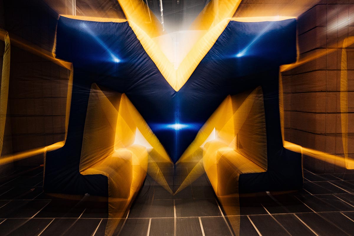 Inflated large block letter "M" in blue and yellow with a motion blur effect.