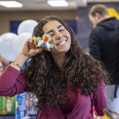 A person with long, curly hair holds a small, colorful object up to their face, standing in a busy indoor setting with balloons and people in the background.