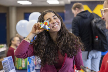 A person with long, curly hair holds a small, colorful object up to their face, standing in a busy indoor setting with balloons and people in the background.