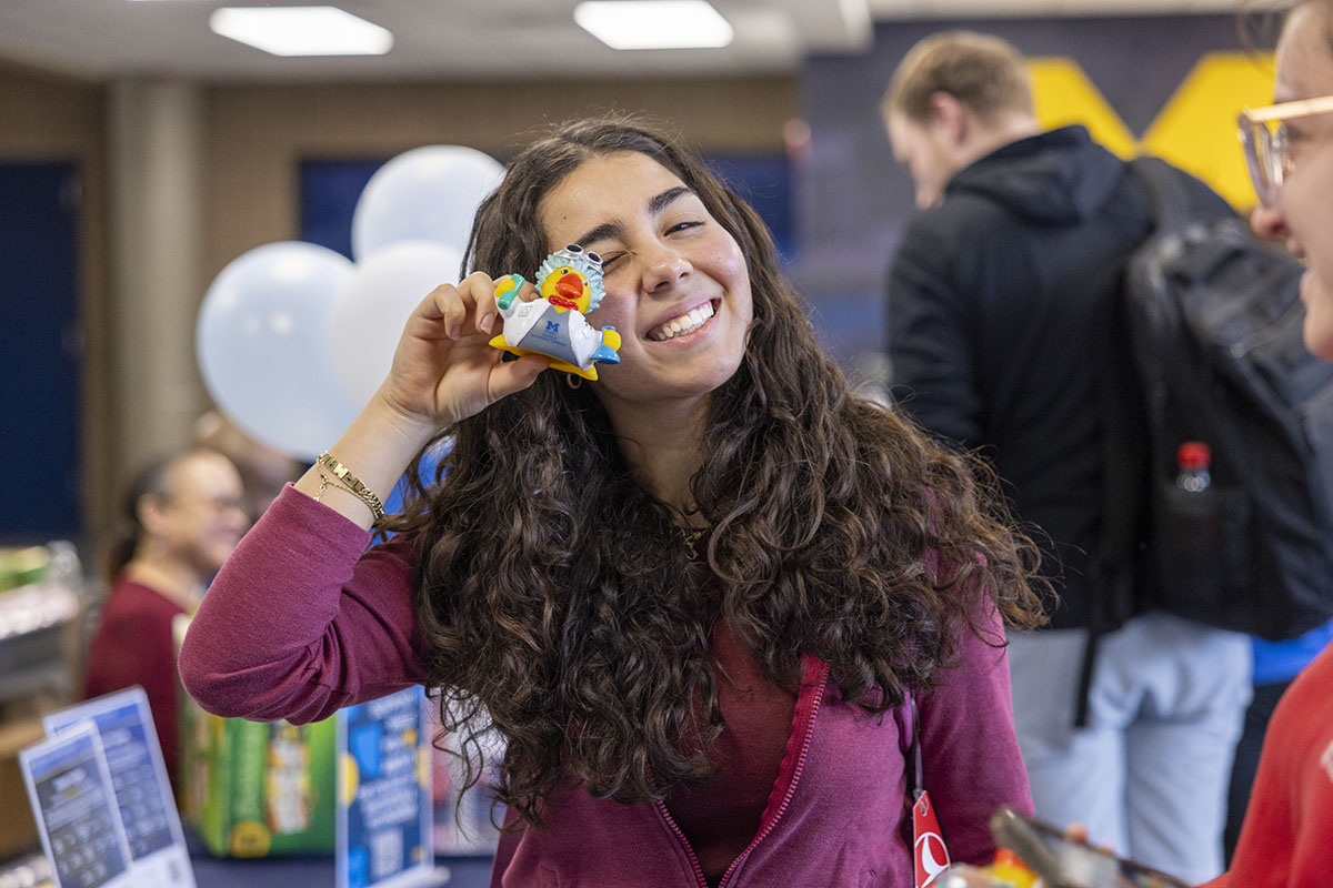 A person with long, curly hair holds a small, colorful object up to their face, standing in a busy indoor setting with balloons and people in the background.