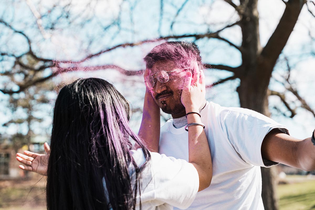 Two people outdoors, one with long dark hair holding the other's face while colored powder is thrown in the air.