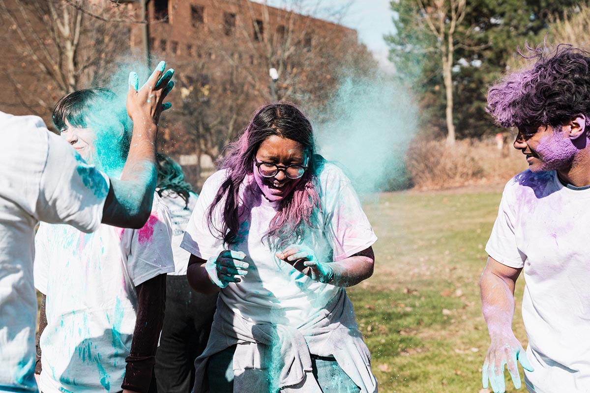 A group of people outdoors wearing white shirts, throwing and covered in colored powder during a color run or festival.