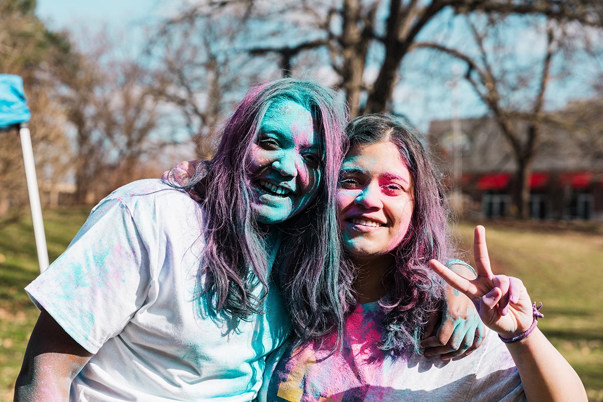 Two people with colorful powder on their hair and clothes pose outdoors, one making a peace sign with their hand.
