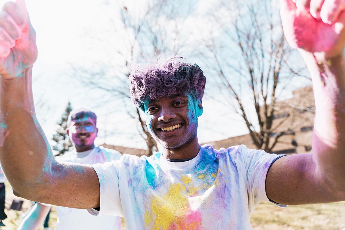 Two people outdoors wearing white shirts covered in colorful powder, with trees and a building in the background.