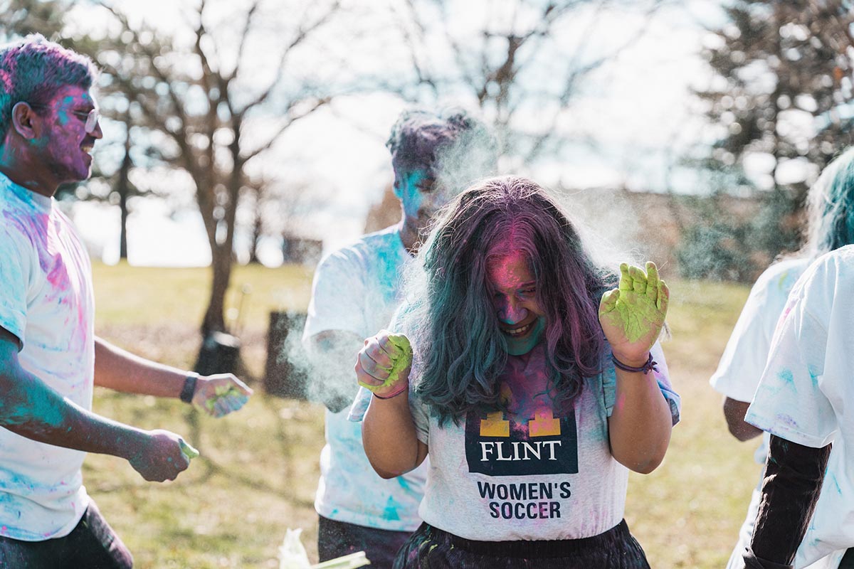 Group of people outdoors throwing colored powder, one person wearing a Flint Women's Soccer shirt with green powder on their hands.