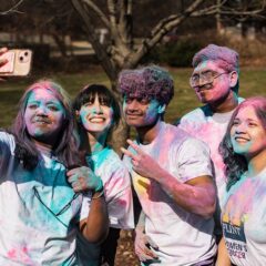 Group of five people covered in colorful powder taking a selfie outdoors in a park.