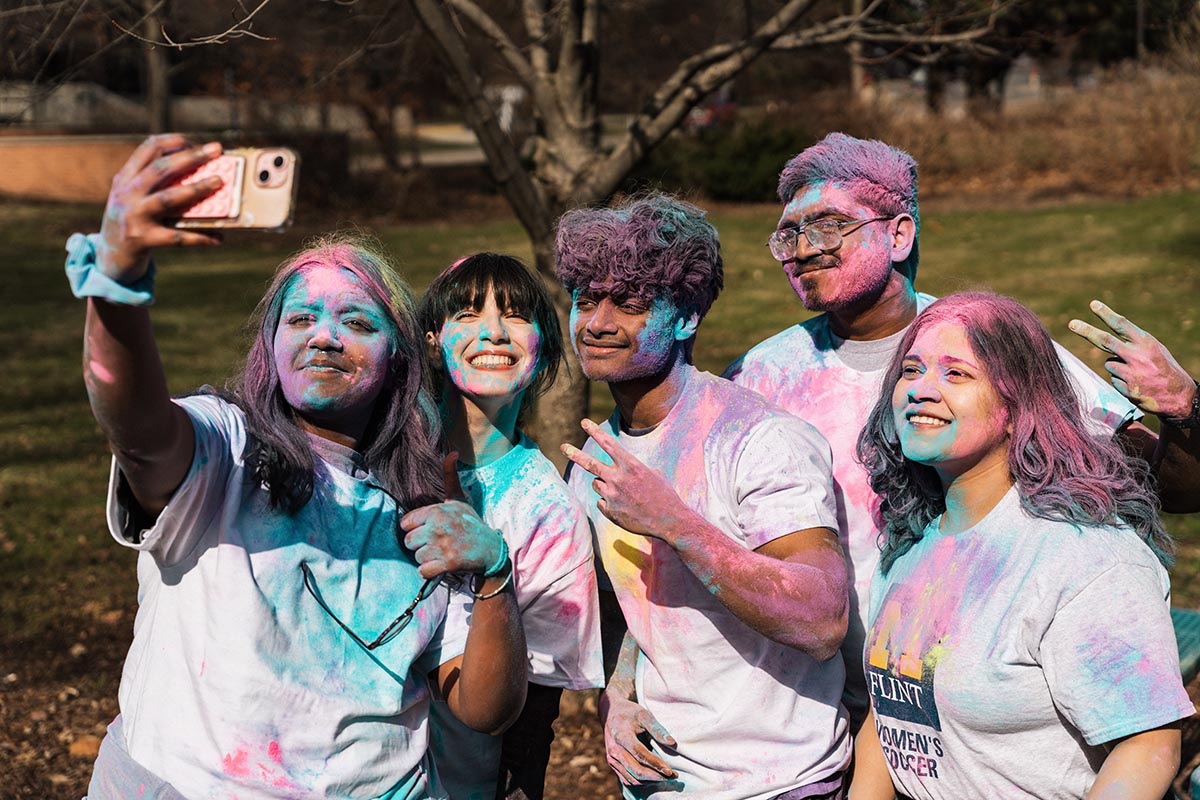 Group of five people covered in colorful powder taking a selfie outdoors in a park.