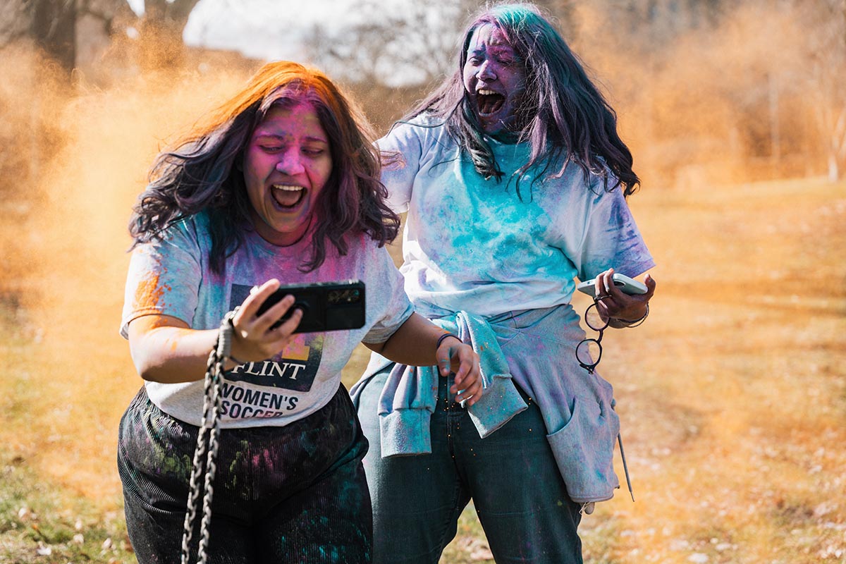 Two people covered in colorful powder, one holding a phone taking a selfie outdoors on a sunny day.