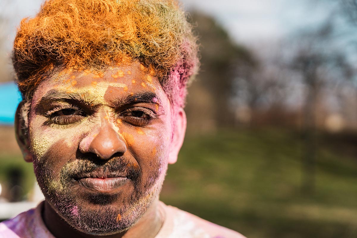 Person with curly hair dyed orange and pink, wearing a tie-dye shirt, outdoors with blurred trees and grass in the background.