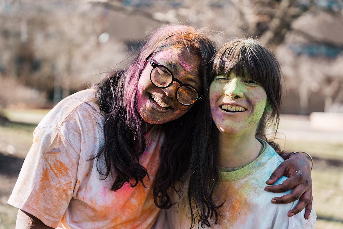 Two people with colorful powder on their white shirts, outdoors with one person's arm around the other.