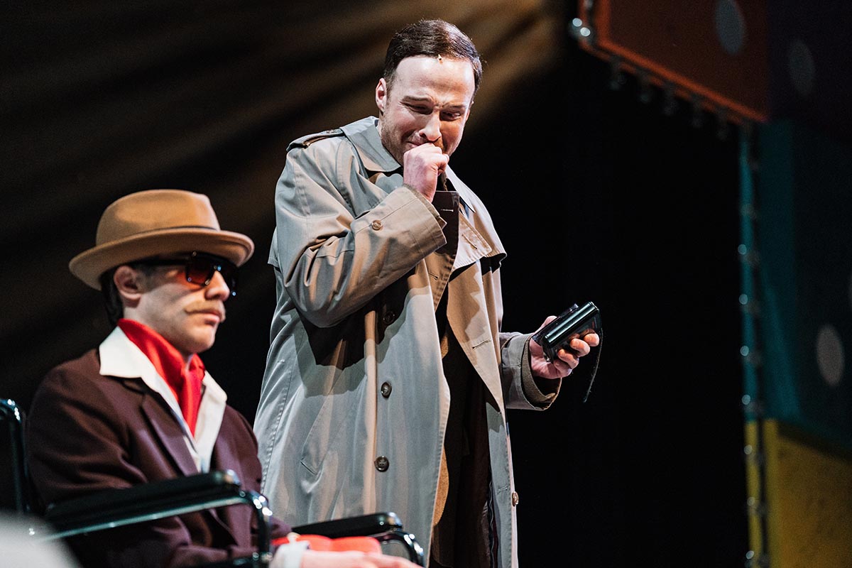 FLINT_Lucky Stiff 2026_0012 Two men on stage, one seated wearing a brown suit, red scarf, and tan hat, the other standing in a beige trench coat holding a small black device.