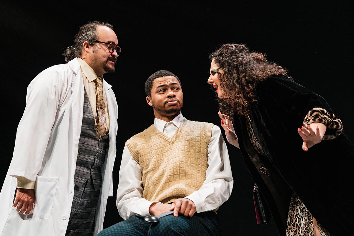 FLINT_Lucky Stiff 2026_0013 Three people engaged in a conversation, one wearing a white lab coat, another in a beige sweater vest, and the third in a black coat with leopard print cuffs.