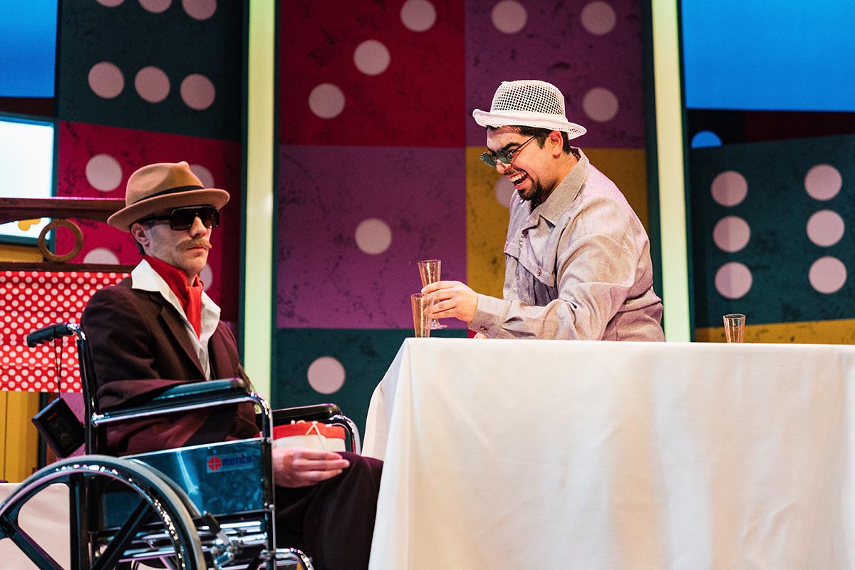 FLINT_Lucky Stiff 2026_0019 Two people in hats are seated at a table with a white tablecloth, one in a wheelchair and the other holding a glass, against a colorful polka-dot background.