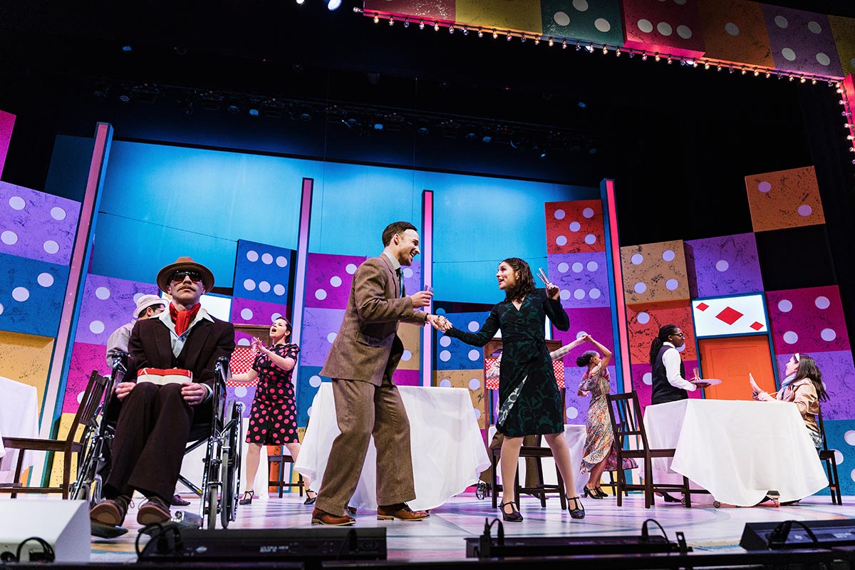 FLINT_Lucky Stiff 2026_0024 Actors performing on a colorful stage set decorated with large domino and playing card designs, with tables and chairs arranged like a restaurant.