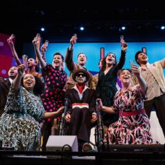 Group of people on stage raising glasses in a celebratory toast, with colorful set design in the background.